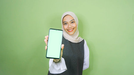 young asian woman wearing hijab, showing copy space on her phone screen. Isolated over green backgroundの写真素材