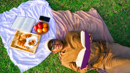 Young asian man reading a book on a picnic with bread, croissants and fruitの写真素材
