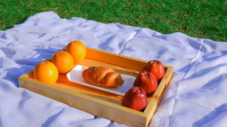 Orange, apples and croissant in a wooden tray on the grass