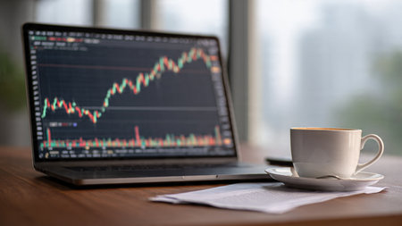 A wooden office desk with economic indicator charts glowing on a laptop beside a coffee cup.の写真素材