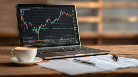 The laptop displays market fluctuations next to printed reports and a warm coffee on a smooth wooden desk.の写真素材