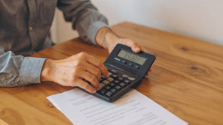 A person using a calculator on a wooden desk with a paper next to it.の写真素材