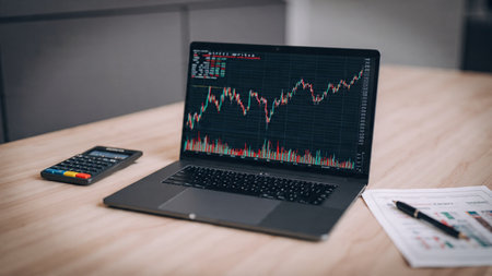 A laptop full of forex, crypto, and stock charts stands beside reports and a coffee mug.の写真素材