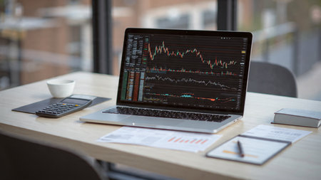Laptop displaying real-time economic charts beside financial paperwork and a coffee cup.の写真素材