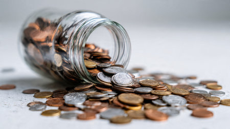 Modern savings presentation featuring a transparent jar angled slightly with coins pooled around it, commercial-safe.の写真素材