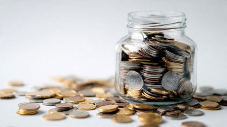 Clear jar placed on a white surface surrounded by small coin stacks representing gradual wealth...