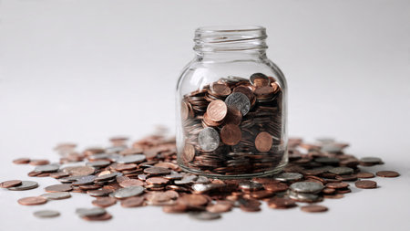 High-key scene showing a jar slightly tilted as coins spill gently across a matte white surface,...
