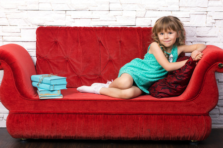  little beautiful girl in fashion dress with long blond curl learn on red cough, near learn pile of...
