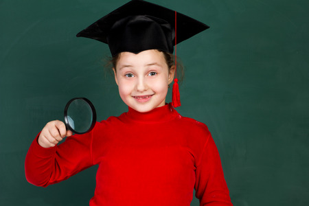 little joyful schoolgirl holding magnifying glass and  trying university hat on chalkboard...
