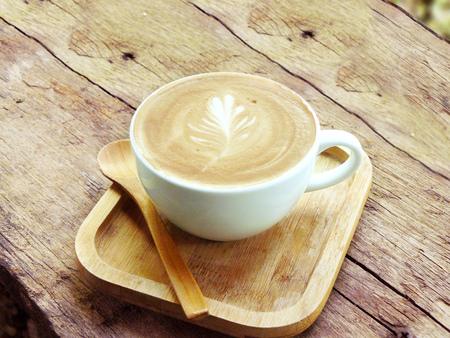 Hot cup of coffee cappuccino decorated leaf line by white foam in a white cup with wooden spoon and plate on natural wooden table.の写真素材