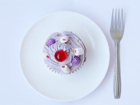 Snow frost cupcake. Vanilla cake in paper cup decorated with frosting creamy, colorful sugar, red jelly ball and marshmallow. Close Up top view in a white plate with fork isolated on white background.の写真素材