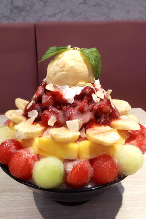Delicious Asia-Asian eating food, Bing su: Korean shaved milk ice dessert with sweet toppings, various mix fruit, strawberry jam and vanilla ice-cream in black ceramic bowl on the table, close upの写真素材