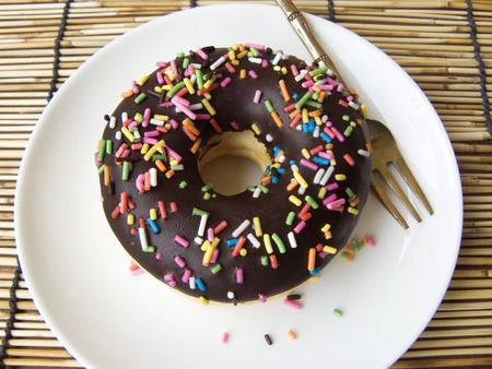 Close up top view of Chocolate Donut decorated with colorful sugar topping on a white plate and fork on Rattan palm mat background.の写真素材