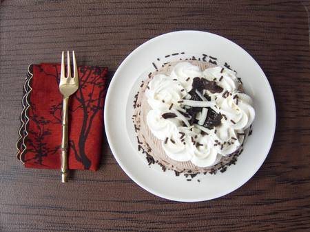 Delicious sweet Chocolate Cream Cake. The chocolate cake decorated with white-black chocolate chip,white cream,dark sugar color on white plate and wooden tray background with brass fork and red napkinの写真素材