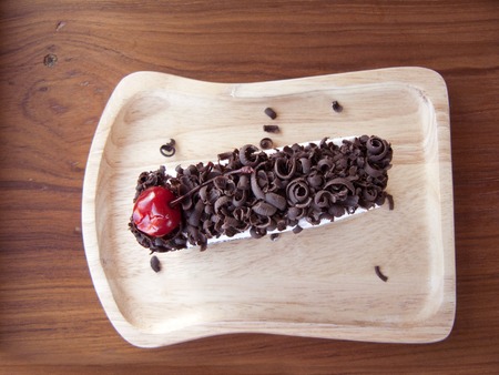 Close up top view of Black Forest Roll Cake, Delicious the chocolate-rum roll cake decorated with cream, red cherry and chocolate sugar color on a wooden plate on wooden table background.の写真素材