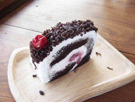 Close up top front of Black Forest Roll Cake, Delicious the chocolate-rum roll cake decorated with cream, red cherry and chocolate sugar color on a wooden plate on wooden table background.の写真素材