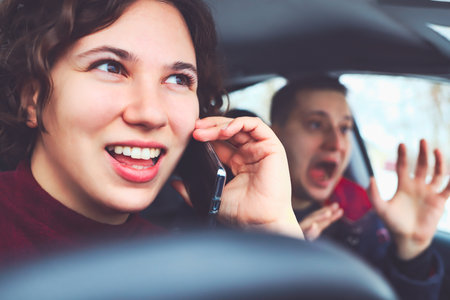Happy curly haired girl talking on mobile phone while driving car. man in next seat is terrified of...