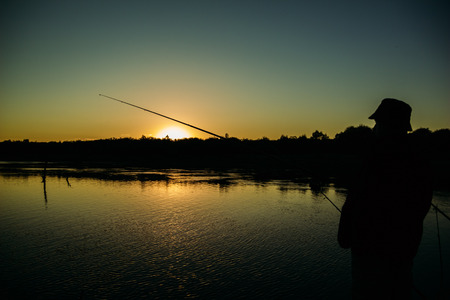 Male angler stands at the river with a fishing rod on the sunset backgroundの写真素材