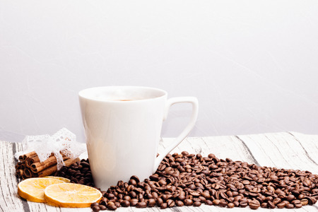 White cup lying beside cinnamon sticks and dry lemon and a scattering of coffee beans on a white backgroundの写真素材