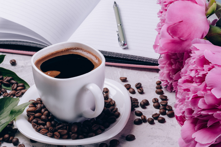 Coffee in a white cup on the table against the background with peonies and a notebookの写真素材