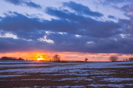 Sunset with orange sun in the clouds over snowy rural fieldsの写真素材