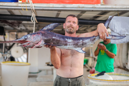 Bali, Island Crete, Greece, - June 30, 2016: Man is a fisherman carries a big fish (sawfish) after successful fish catch from the fishing boat.のeditorial素材