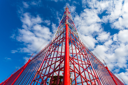 Telecommunication tower with panel antennas and radio antennas and satellite dishes for mobile communications (2G, 3G, 4G, 5G) with red fence around tower against blue with cloudsの写真素材