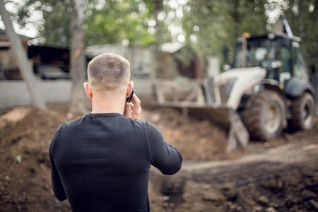 Young man speaks by phone on the construction site when the modern excavator performs excavation work on the construction site during reconstruction placeの写真素材