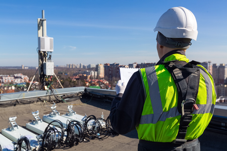 Professional industrial climber in helmet and uniform reads technical documentation and  antennas of GSM  DCS UMTS LTE bands, outdoor radio units  on the roof. Working process of upgrading telecommunication equipment.の写真素材