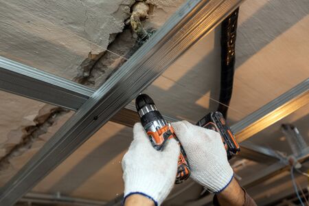 Worker with protective gloves is drilling by perforator the ceiling for installing metall frames  in...