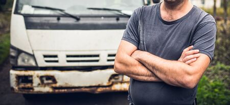 Male driver in old-fashioned t-shirt has crossed hands and stays near his old rusty truck on the parking place.の写真素材