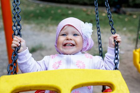 Adorable smiling baby girl with kerchief enjoying a swing ride on a playground in a park on a nice sunny summer dayの写真素材