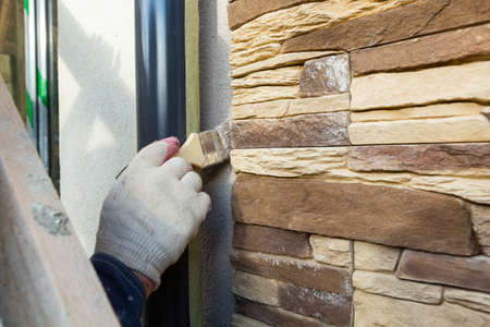 Worker in protective gloves is painting by the brush the facade house that is under construction,...