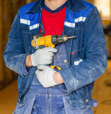 Worker in dirty uniform and protective gloves with drill in his hand in the room of apartment that is under construction, remodeling, renovation, extension, overhaul, restoration and reconstruction.の写真素材