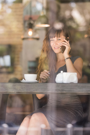 Young woman  sitting  by the window at the bar counter in coffee shop, and talking on her cell phone  の写真素材