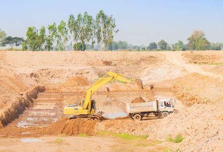 The truck is loading soil from backhoe at the bottom of the pond.の写真素材