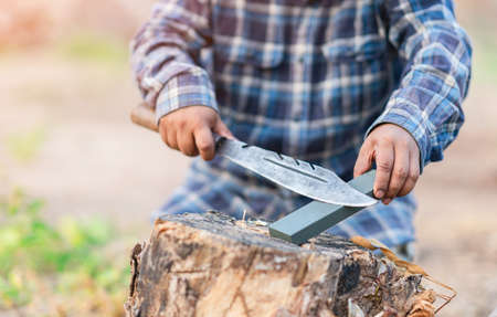 A man sharpening a knife hiking on a tree stump out of focus background.の写真素材