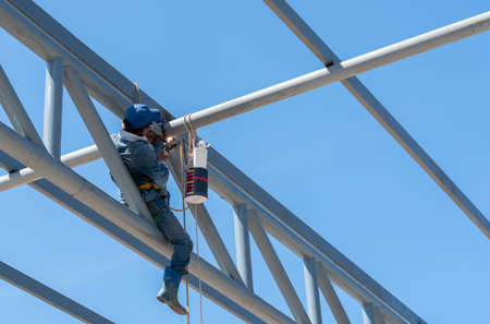 Welders working at dangerous heights on grey painted steel roof truss. Welding of steel is very technically demanding and is only professional. Bright sky backgroundの写真素材