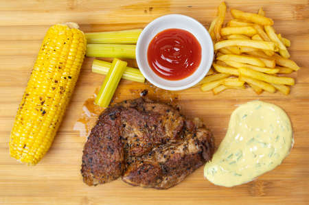 Danish-style pork steak, roasted corn, french fries, pickles and ketchup are placed on a wooden cutting board.の写真素材