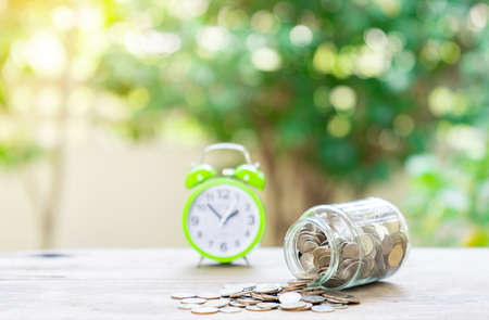 Pension planning save money for retirement.Concept coin money in glass jar and alarm clock on blurred green background.の写真素材