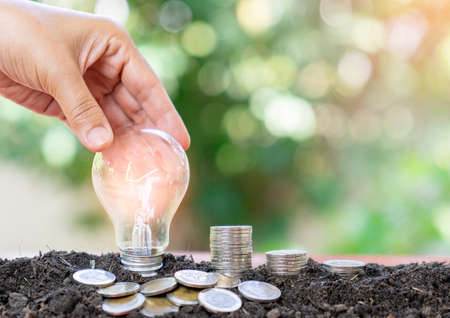 hand holding and putting light bulb on pile of coins on soil for saving energy and money conceptの写真素材