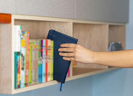 Male left hand choosing and picking blue book in home book shelf. Education research and self learning at home life conceptsの写真素材