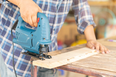 A carpenter uses an electric  jigsaw to cut wooden bars on the table, out of focus background.の写真素材