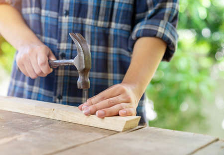 Hammer in hold carpenter hand hit nail on wood bar, out of focus background.の写真素材