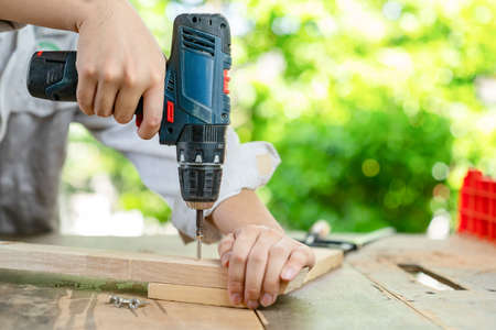 Close up hand hold cordless screwdriver tighten screw on wood plank, out of focus background.の写真素材