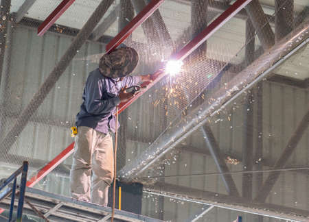 A man welder working at height with spark under roof in factory.