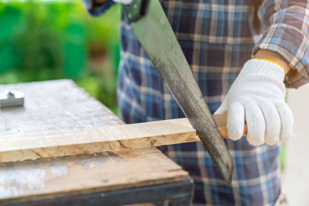 The carpenter ware white gloves use hand saw cutting wood board in shop.