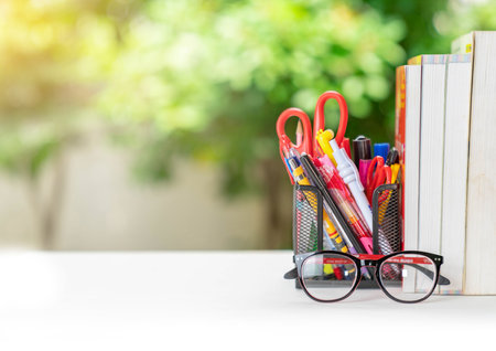 A stack of books, glasses, stationery on a table and a out of focus green tree backgroundの写真素材