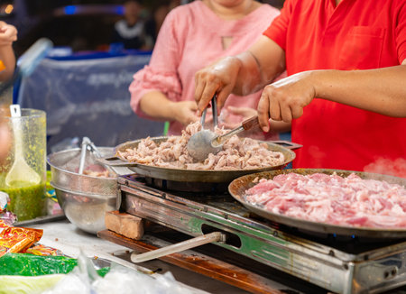 The vendor is grilling meat in pan on a stove in street food market.