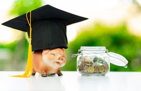 A piggy bank wearing a graduation cap with coins in a jar on a blurred background.の写真素材
